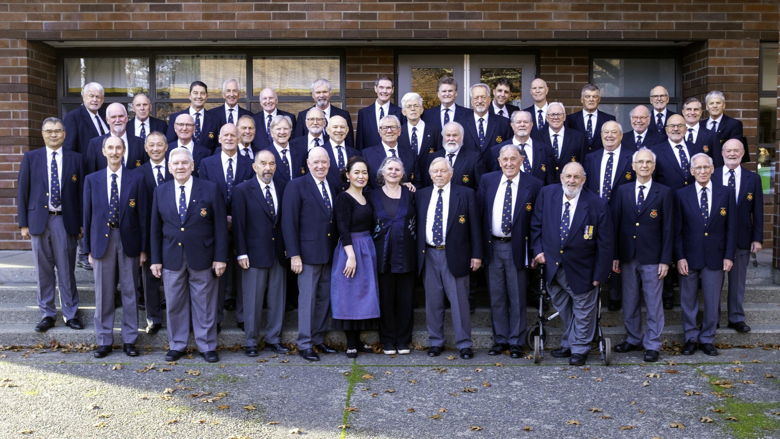 A photo of the Vancouver Welsh Men's Choir, including the Choir Director, Alison Nixon, and pianist, Miri Lee. The photo includes a large group of men standing outdoors, all wearing blue blazers. Alison and Miri are in the front, dressed in formal performance wear.