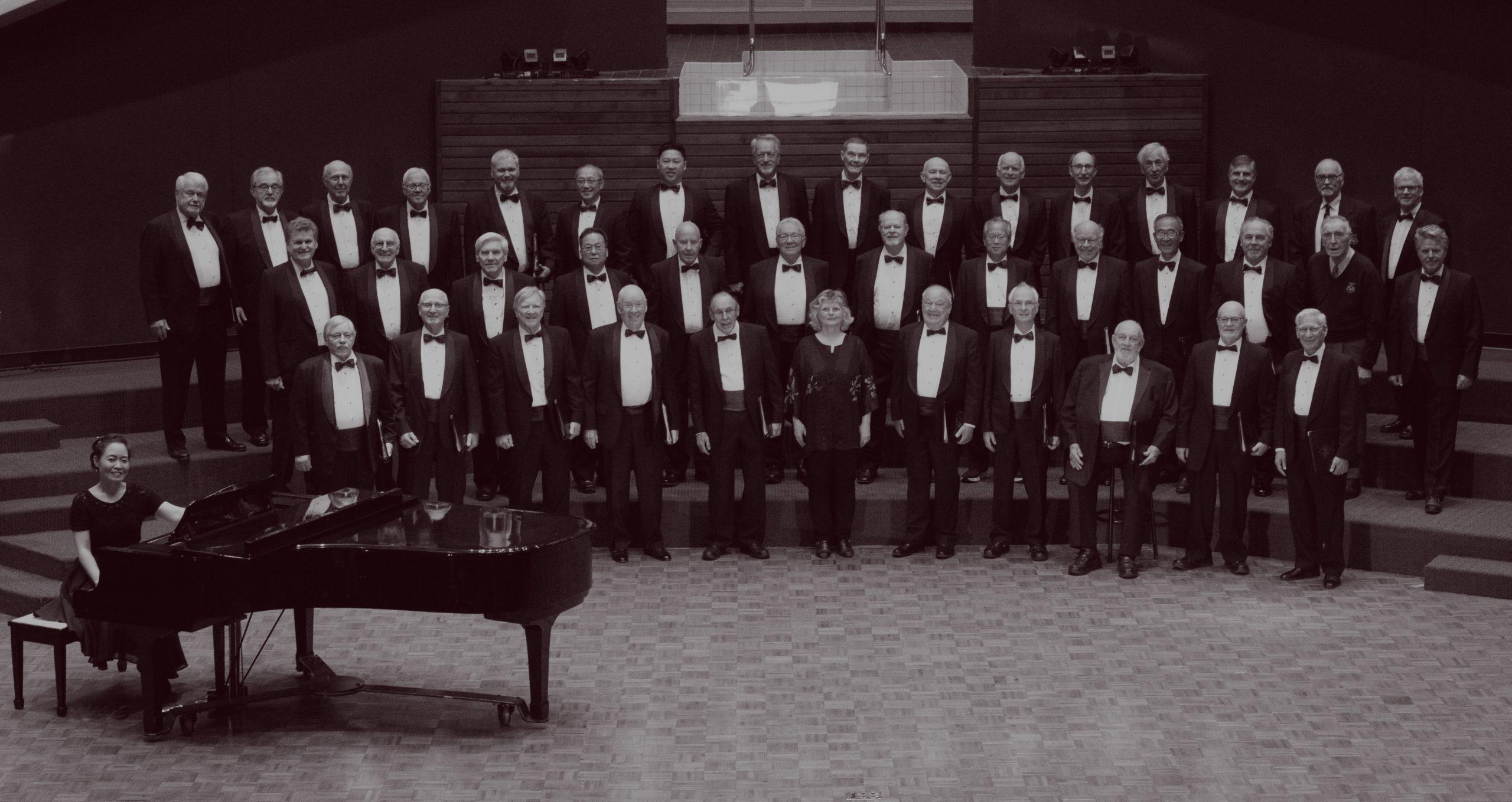 The Vancouver Welsh Men's Choir on stage at a performance. Includes approximately 50 men in tuxedos on stage, with a piano off to the side and Miri Lee sitting at the piano.